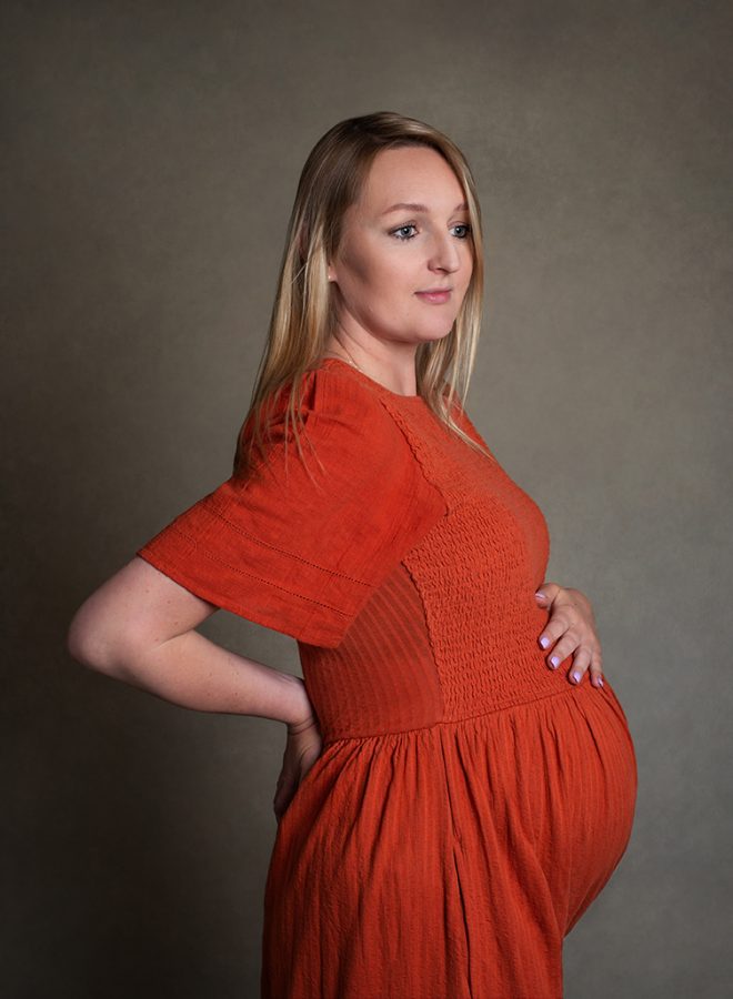 A white woman photographed side on to camera. She has long blonde hair and wears a bright orange dress with elbow length sleeves. She's pregnant and rests one had on top of bump and the other hand is on her back. She looks off camera